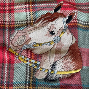 May include: An embroidered patch of a horse head in profile, featuring brown and white fur, wearing a yellow and silver bridle with decorative metal accents, set against a background of red, green, and white plaid fabric with thin yellow lines.