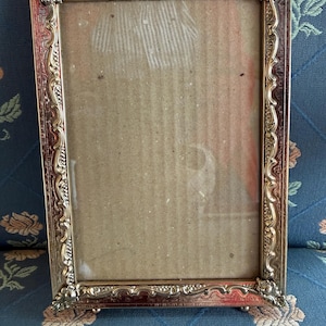 May include: An ornate, gold-coloured rectangular picture frame. The frame features a detailed, scrolling design and decorative corner accents. The frame is empty, with a cardboard backing visible through the glass.