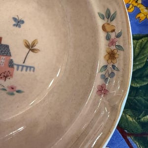 May include: A ceramic bowl with a speckled beige finish featuring a painted design of a small pink house with a blue roof, a blue butterfly, and a floral garland along the rim consisting of yellow, pink, and blue flowers with green leaves.
