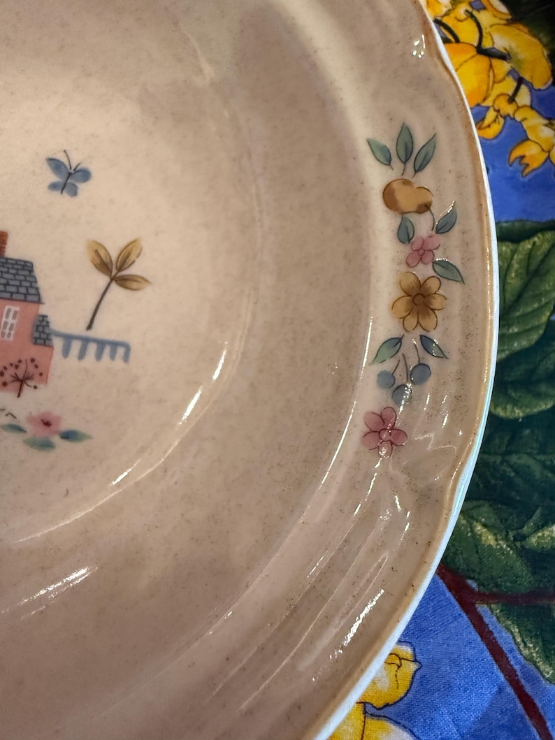 May include: A ceramic bowl with a speckled beige finish featuring a painted design of a small pink house with a blue roof, a blue butterfly, and a floral garland along the rim consisting of yellow, pink, and blue flowers with green leaves.