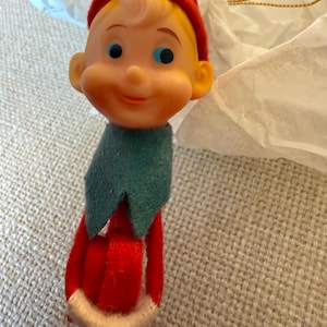 Puede incluir: Adorno navideño vintage con un elfo sonriente con sombrero y atuendo rojos, cuello verde y ribete crema. El elfo tiene ojos azules y cabello rubio. El adorno está hecho de fieltro y plástico.