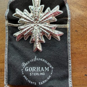 May include: A silver snowflake ornament with intricate geometric designs rests on a dark gray Pacific Silvercloth bag. The bag features the Gorham Sterling logo and text that reads "Prevents Tarnish."