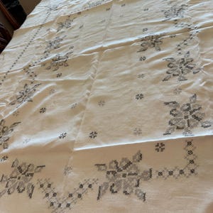 May include: Cream-colored tablecloth with a floral cross-stitch pattern in shades of gray. The design features stylized flowers and geometric shapes, creating a vintage aesthetic. The fabric appears to be linen or a similar natural material.