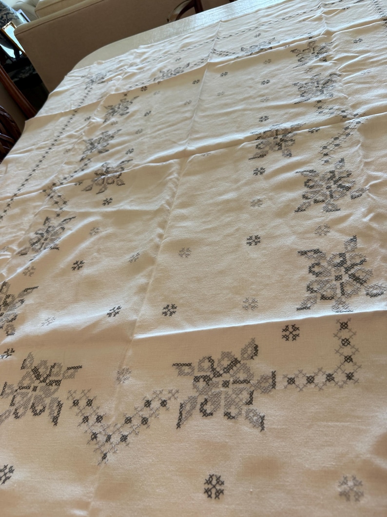 May include: Cream-colored tablecloth with a floral cross-stitch pattern in shades of gray. The design features stylized flowers and geometric shapes, creating a vintage aesthetic. The fabric appears to be linen or a similar natural material.