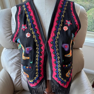 Pode incluir: Colete preto bordado com franjas. O colete apresenta detalhes coloridos em vermelho, azul, amarelo e verde. O bordado inclui estrelas e corações. A peça de roupa é exibida em um manequim.
