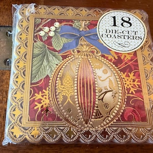 May include: A package of 18 die-cut coasters. Each coaster features a gold-framed design with a red background, a gold ornament, a blue bow, and white berries. The coasters are in a clear plastic package.