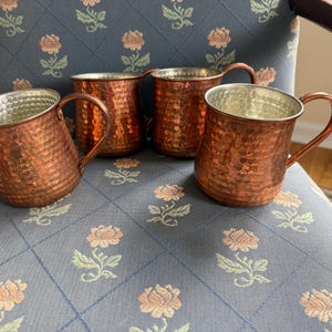 Puede incluir: Cuatro tazas de cobre con exteriores martillados e interiores plateados sobre una silla con estampado floral. Cada taza tiene un asa curvada. El color cobre es un marrón rojizo cálido.