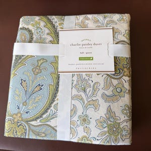 Può includere: Un copripiumino Charlie Paisley piegato in una confezione, con un motivo paisley floreale nei toni del verde, blu e beige. La confezione è fissata con una fascia bianca e include un'etichetta del prodotto con la scritta "full - queen" e "organic".