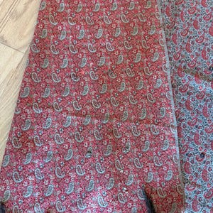 May include: A red paisley patterned textile with a black fringe. The fabric features a repeating design of small, teardrop-shaped motifs in shades of gray and white against a red background. The textile is folded, revealing the pattern.