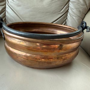 May include: A round, hammered copper basin featuring a wide, dark iron handle that arches over the top. The copper vessel has a polished, metallic finish with visible horizontal ridges along its exterior body and two sturdy iron side attachments.