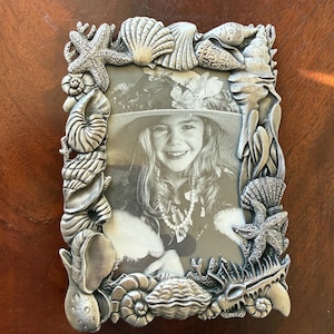 Könnte beinhalten: Silberfarbener Bilderrahmen mit detailliertem Muscheldesign. Der rechteckige Rahmen zeigt Seesterne, Muscheln und Korallen. Er enthält ein Schwarz-Weiß-Foto einer lächelnden Person.