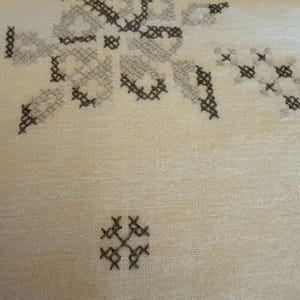 May include: Close-up of a cream-colored fabric with embroidered cross-stitch patterns in shades of gray and brown. The design features floral and geometric shapes, creating a textured surface.