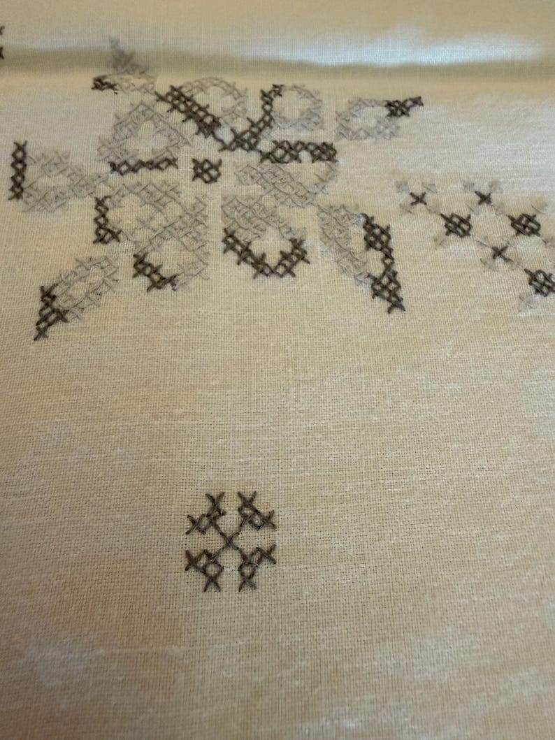 May include: Close-up of a cream-colored fabric with embroidered cross-stitch patterns in shades of gray and brown. The design features floral and geometric shapes, creating a textured surface.