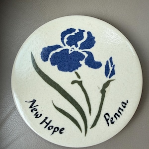 Puede incluir: Un posavasos redondo de cerámica blanquecina con un diseño de flor de iris azul. La flor está centrada, con tallos y hojas verdes. Las palabras "New Hope Penna." están escritas en negro alrededor del borde.