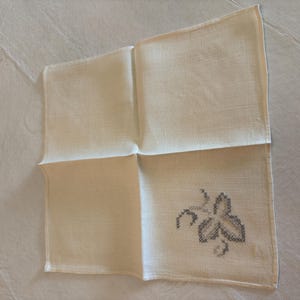 May include: Cream-colored linen handkerchief with a cross-stitched leaf design in shades of gray. The square handkerchief is folded, showcasing the embroidered detail in one corner. A vintage textile.