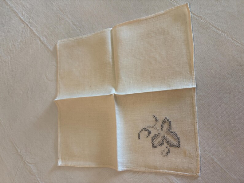 May include: Cream-colored linen handkerchief with a cross-stitched leaf design in shades of gray. The square handkerchief is folded, showcasing the embroidered detail in one corner. A vintage textile.