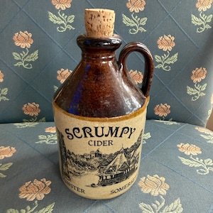 May include: A brown ceramic jug with a handle and a cork stopper. The jug features a label with the words "SCRUMPY CIDER" and a black and white illustration of a building and landscape. The jug is sitting on a blue floral patterned surface.