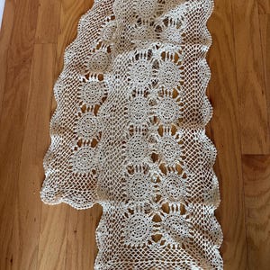 May include: An off-white rectangular crochet table runner featuring a repeating circular floral motif pattern. The piece is constructed with intricate openwork stitching and has a scalloped edge along the entire perimeter of the textile.