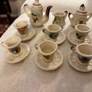 Könnte beinhalten: Ein Miniatur-Teeservice im Vintage-Stil mit sechs Tassen und Untertassen, zwei Teekannen und einem Sahnekännchen. Das Keramik-Set zeigt Illustrationen von Zeichentrickfiguren in verschiedenen Farben. Die Tassen und Untertassen haben einen Durchmesser von ca. 4 cm.