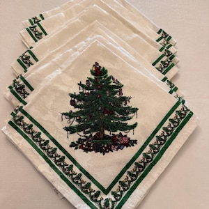 May include: A stack of square white cloth napkins featuring a central embroidered Christmas tree decorated with ornaments and gifts. Each napkin has a green border with a repeating holly leaf and berry pattern along the edges.