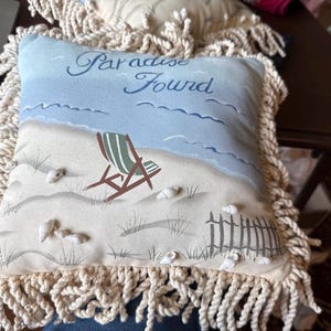 May include: A decorative pillow with a beach scene. The pillow features a painted beach with a blue sky, ocean, and a beach chair. The words "Paradise Found" are written across the top. The pillow is trimmed with a cream-colored fringe.