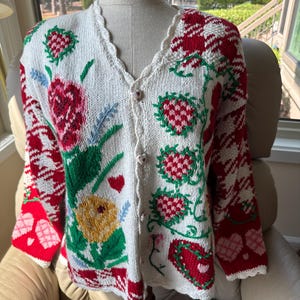 Peut inclure: Un gilet en tricot couleur crème avec un col en V à bords festonnés et des boutons en forme de cœur. Le devant présente des motifs floraux et de fraises colorés en rouge, vert et jaune, tandis que les manches arborent un motif rouge et blanc.