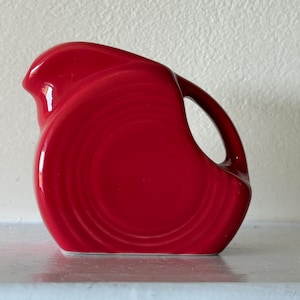May include: A bright red ceramic jug with a curved handle and a spout. The jug has a glossy finish and a smooth, rounded body. The design features concentric circles on the front, adding visual interest to the piece.