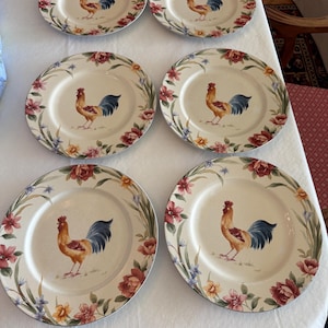 May include: A set of six round ceramic dinner plates featuring a central illustration of a rooster with brown, red, and blue feathers. The rim of each plate is decorated with a floral pattern consisting of red, yellow, and blue flowers and green leaves.