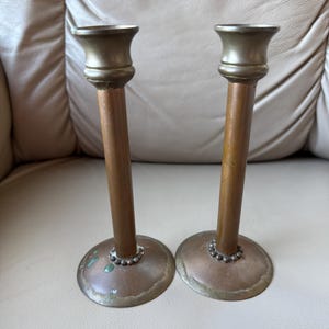 May include: Two antique copper candlesticks with a tarnished patina. The candlesticks have a round base, a long, slender stem, and a flared top. The base is decorated with a ring of small, round beads. The candlesticks are displayed on a neutral background.