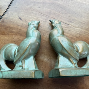 May include: A pair of metallic bookends shaped like roosters, featuring a weathered green and gold finish. Each rooster stands on a rectangular base, with stylized feathers and an upright posture, designed to support books on a shelf or flat surface.