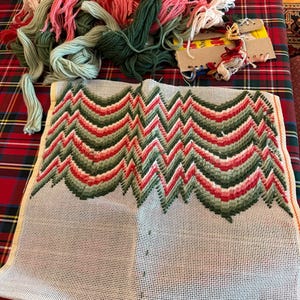 May include: A needlepoint project in progress, featuring a canvas with a zig-zag pattern in red, green, and pink. Bundles of yarn in matching colors are scattered nearby. The project is on a red and green plaid surface.