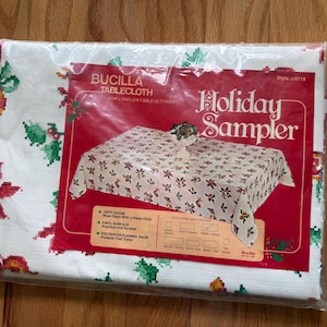Könnte beinhalten: Eine verpackte Bucilla Holiday Sampler Tischdecke. Die weiße Tischdecke hat ein Muster aus roten und grünen floralen Designs. Die Verpackung ist rot und weiß mit den Worten "Holiday Sampler" und "Bucilla Tablecloth".
