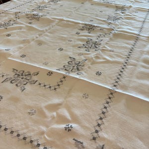 May include: Cream-colored tablecloth with embroidered floral and geometric patterns in gray. The tablecloth is rectangular and covers a large table, showcasing detailed needlework and a classic design.