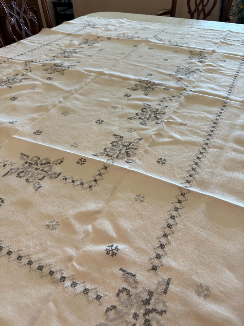 May include: Cream-colored tablecloth with embroidered floral and geometric patterns in gray. The tablecloth is rectangular and covers a large table, showcasing detailed needlework and a classic design.