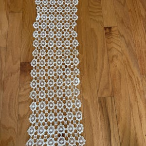 May include: Cream-colored, open-weave lace table runner. The runner features a repeating floral and geometric pattern. The edges are scalloped. The runner is displayed on a hardwood floor.