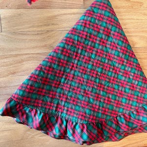 May include: A quilted tree skirt featuring a red and green plaid pattern with a ruffled edge. The fabric has a stitched diamond texture throughout the surface, creating a soft, layered appearance with a circular shape and a gathered border.