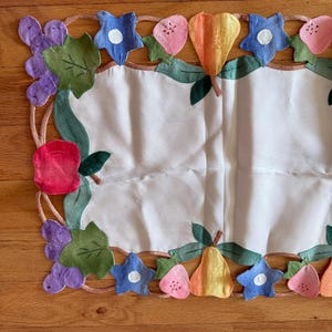 May include: A rectangular white fabric placemat featuring a decorative border of appliqued fruit and flowers. The border includes purple grapes, green leaves, red apples, pink strawberries, yellow pears, and blue flowers with white centers.