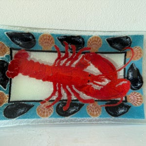 May include: A rectangular glass tray featuring a central white panel with a large, vibrant red lobster illustration. The border is a bright turquoise blue, decorated with alternating black mussel shells and tan scallop shell motifs.