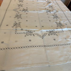 May include: White tablecloth with a floral pattern in shades of gray. The design features stylized flowers and geometric borders, creating a vintage aesthetic. The tablecloth is spread across a table, showcasing its full size and intricate details.