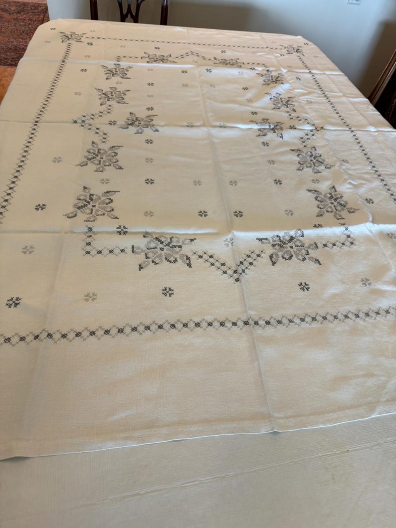 May include: White tablecloth with a floral pattern in shades of gray. The design features stylized flowers and geometric borders, creating a vintage aesthetic. The tablecloth is spread across a table, showcasing its full size and intricate details.