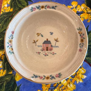May include: A round ceramic bowl with a speckled cream-colored base. The center features a small pink house with a blue roof, surrounded by flowers and a butterfly. The rim is decorated with a repeating pattern of colorful flowers and leaves.