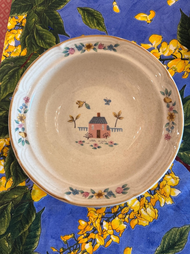 May include: A round ceramic bowl with a speckled cream-colored base. The center features a small pink house with a blue roof, surrounded by flowers and a butterfly. The rim is decorated with a repeating pattern of colorful flowers and leaves.