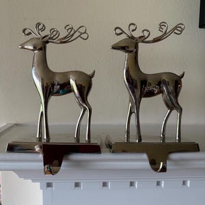 May include: A pair of silver-toned metal stocking holders, each featuring a standing deer figure with stylized, curved antlers. The deer are mounted on rectangular bases, with a hook extending from the front of each base for hanging items.
