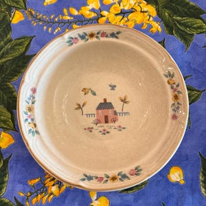 May include: A round, off-white ceramic bowl with a speckled finish. The center features a small pink house illustration with a blue roof, surrounded by tiny flowers and a butterfly. The rim is decorated with a floral pattern of blue and pink.