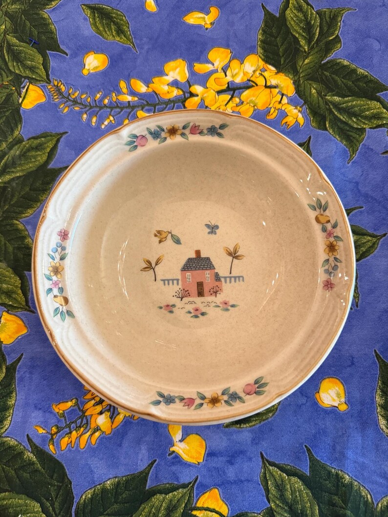 May include: A round, off-white ceramic bowl with a speckled finish. The center features a small pink house illustration with a blue roof, surrounded by tiny flowers and a butterfly. The rim is decorated with a floral pattern of blue and pink.