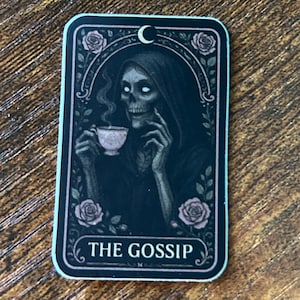 Puede incluir: Adhesivo rectangular con una ilustración gótica de un esqueleto sosteniendo una taza de té. La imagen está enmarcada con rosas y una luna creciente, con las palabras "THE GOSSIP" en la parte inferior. El adhesivo tiene un borde blanco.
