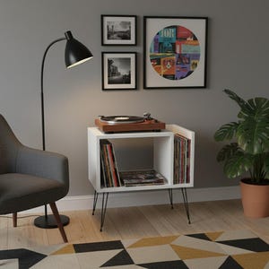 May include: A white record player stand with black hairpin legs. The stand holds a wooden record player, vinyl records, and a collection of albums. A black floor lamp and a gray armchair are nearby. Artwork hangs on the wall.