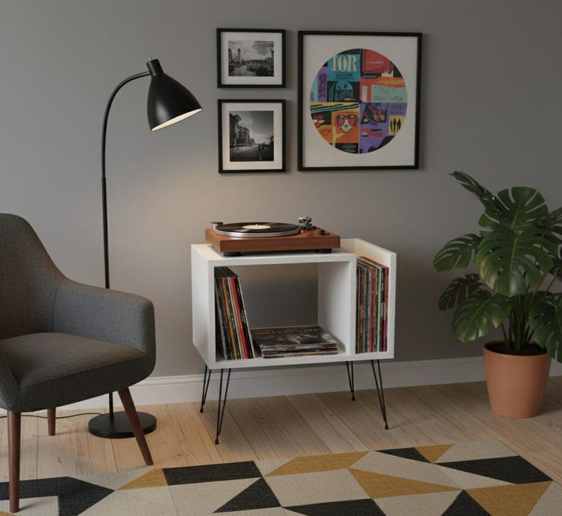 May include: A white record player stand with black hairpin legs. The stand holds a wooden record player, vinyl records, and a collection of albums. A black floor lamp and a gray armchair are nearby. Artwork hangs on the wall.