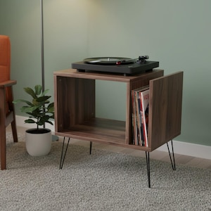 May include: A dark brown, mid-century modern record player stand with black hairpin legs. The stand has an open compartment for storage and a section for vinyl records. A black record player sits on top.