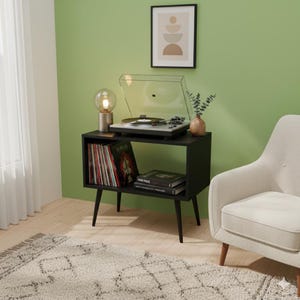 Puede incluir: Un tocadiscos negro sobre un soporte negro con un estante, que contiene discos de vinilo y libros. Una lámpara decorativa y un pequeño jarrón con vegetación están sobre el soporte. La habitación tiene una pared verde y un sillón blanco.